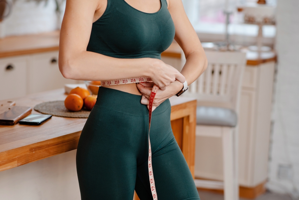 Cropped view of the sporty woman measuring waist with tape at bright kitchen. Weight loss and diet concept.