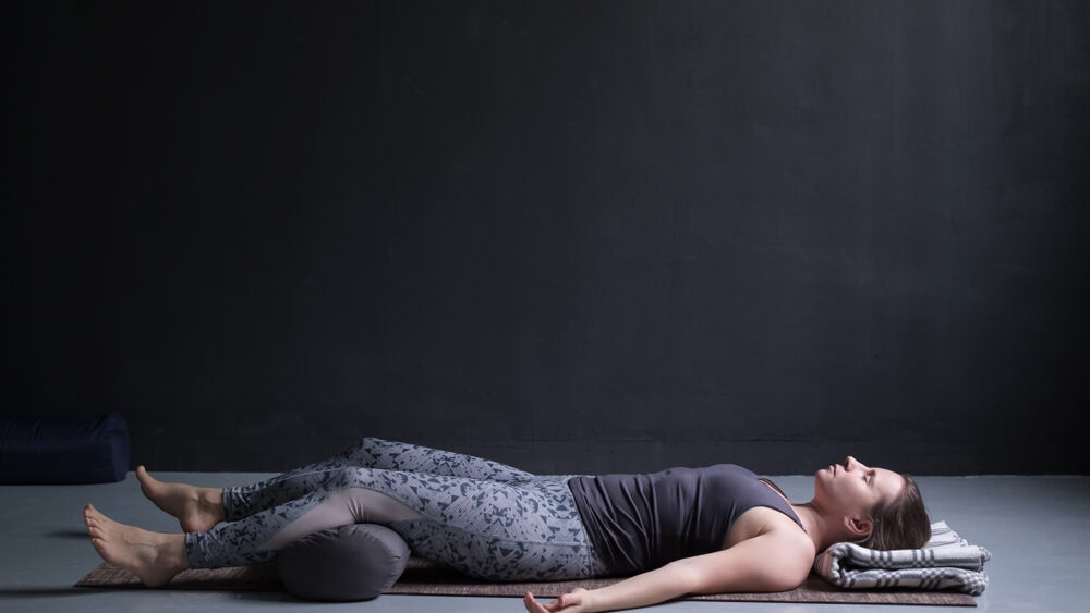 Young fit women lying on the yoga mat with pillow under the legs performing Yog Nidra Asana