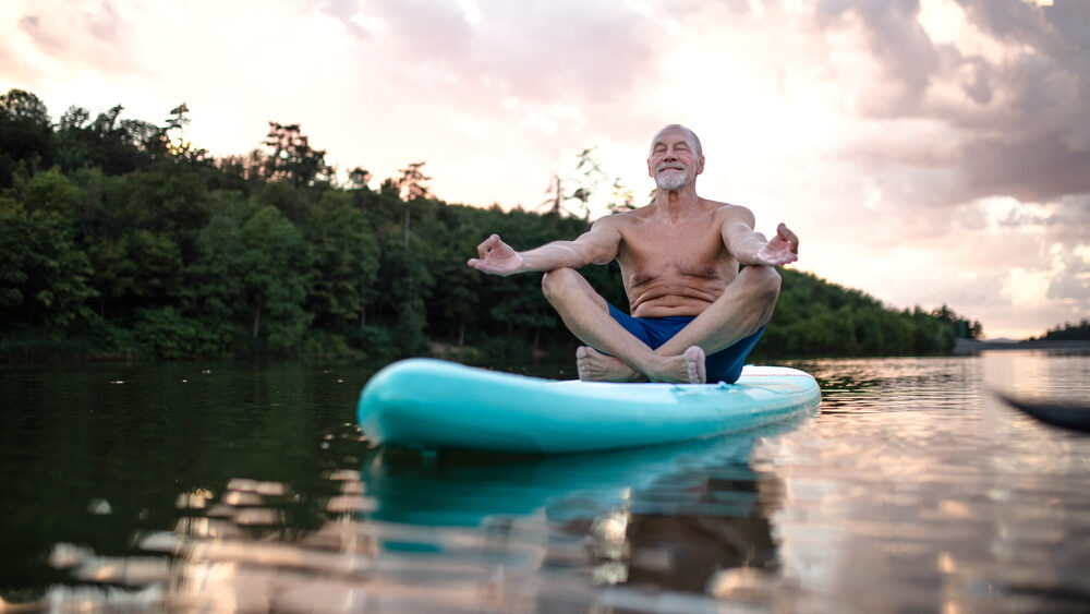 Old fit man balancing on paddle board while meditating/performing yoga in the middle of the river